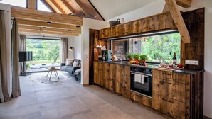 A modern kitchen with wooden cabinetry, a large window with a view of greenery, and a dining area with a sofa and a coffee table.