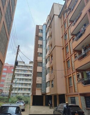 A multi-story residential building with balconies, a central glass elevator shaft, and parked vehicles. - Stylish 1BR retreat in TRM Drive, Nairobi with modern comfort and serene vibes. (Nairobi)