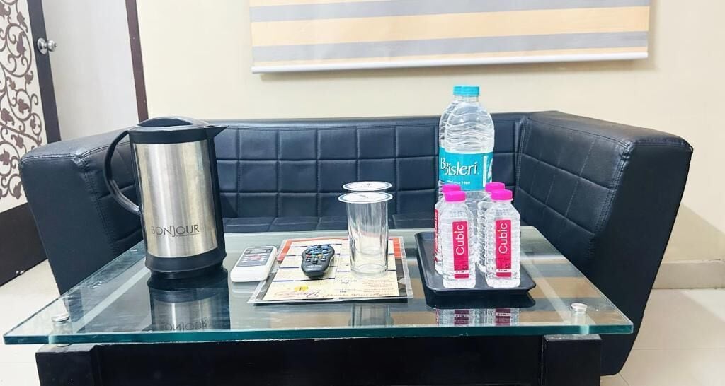 A glass-top table with a black leather couch, a stainless steel coffee maker, a glass, a remote control, a water bottle, and two bottles of Cucumber Cooler.