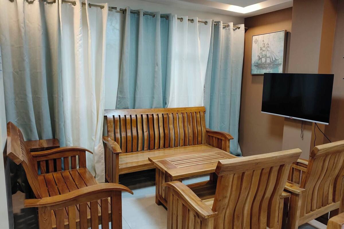 Een kamer met houten meubels, waaronder een bank en stoelen, een klein tafeltje en een aan de muur bevestigde televisie.