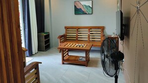 A living room with a wooden sofa, a wooden coffee table, a wall-mounted TV, a standing fan, and a painting on the wall. - MEEK'S VILLAS TRANSIENT HOUSE (Baguio)