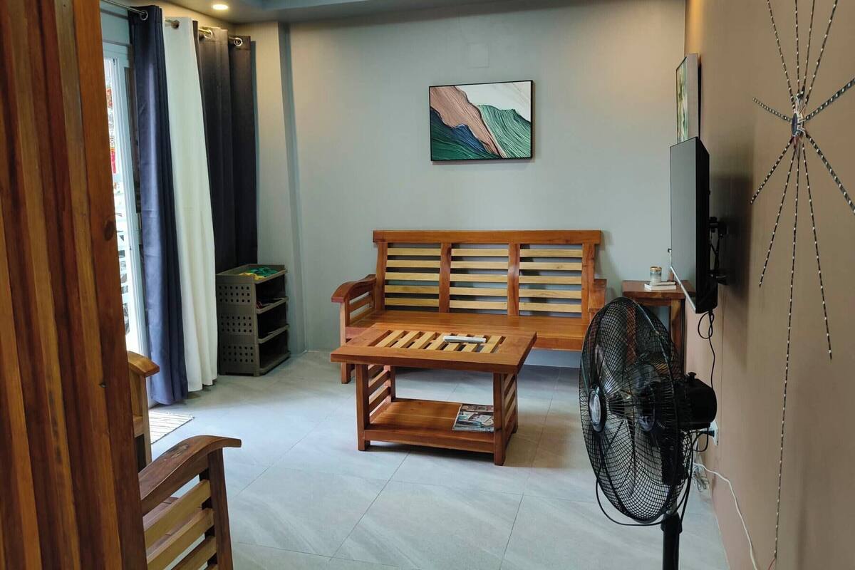 Een woonkamer met een houten bank, een houten salontafel, een aan de muur bevestigde televisie, een ventilator en een schilderij.