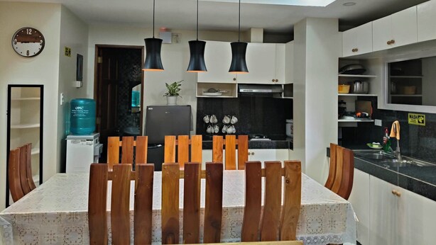 A modern kitchen with white cabinets, a black countertop, and a built-in oven. There is a dining area with a table and chairs, and a clock on the wall. - MEEK'S VILLAS TRANSIENT HOUSE (Baguio)