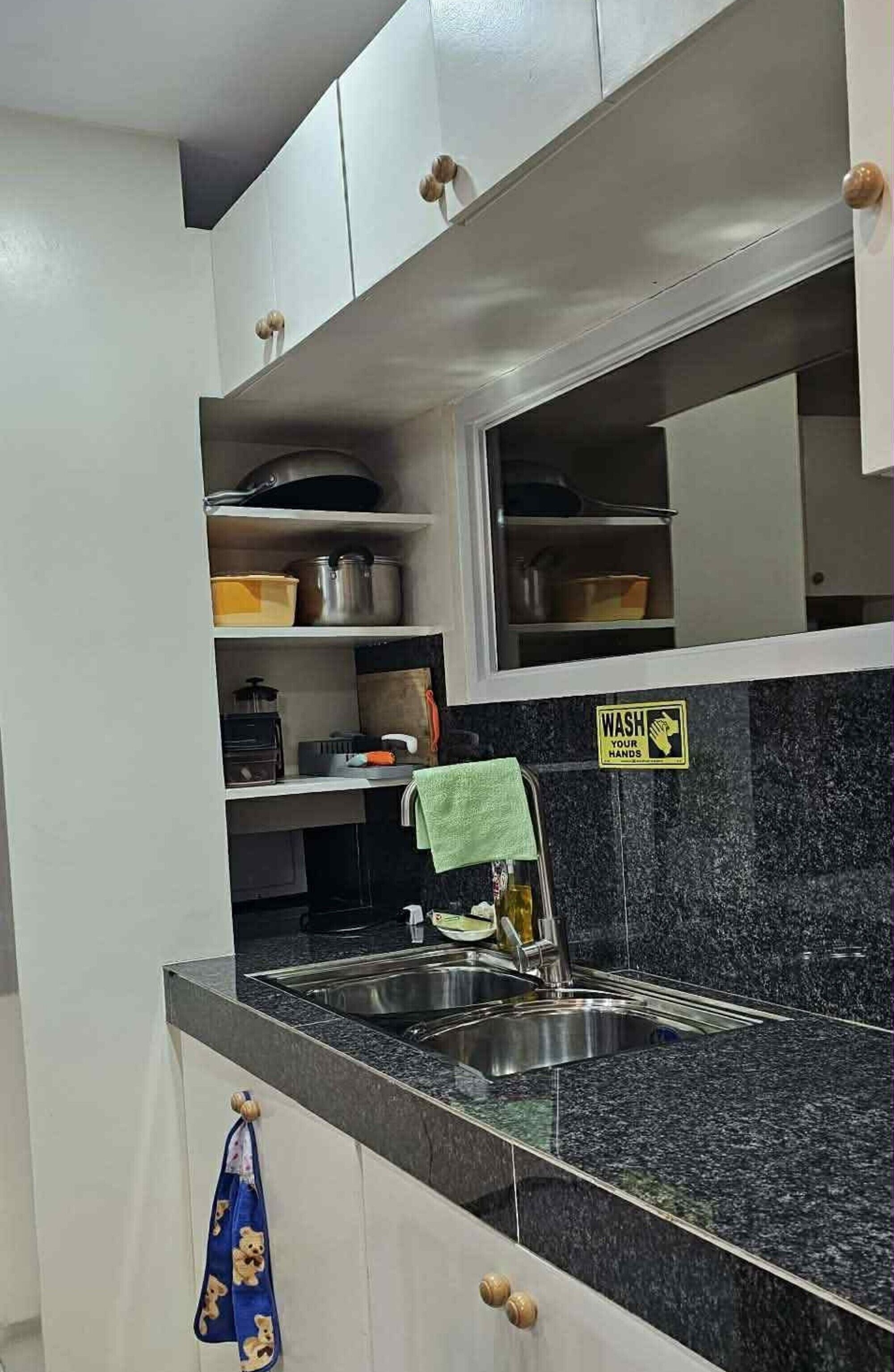 A kitchen with a granite countertop, stainless steel sink, and cabinets with wooden handles. There is a 'WASH YOUR HANDS' sign on the wall.
