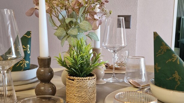 Uma mesa de jantar posta com uma vela, uma planta em vaso, um copo com líquido escuro, um copo transparente e uma embalagem de petiscos.