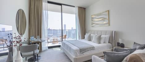 A modern hotel room with a large bed, a sitting area with a sofa and armchair, a dining table with chairs, a large window with curtains, and a city view.