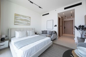 A modern bedroom with a large bed, a sofa, a bedside table, a wall-mounted artwork, and a door leading to another room. - Bloomfields Cozy Stu Binghatti (Dubai)