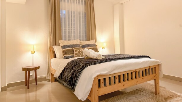 Un dormitorio con cama de madera, dos lámparas de noche, una ventana con cortinas translúcidas y una lámpara en el techo.