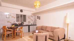 Sala de estar con un sofá color beige, sillas de madera, una mesa de centro, una lámpara y una zona de cocina con salpicadero de azulejos y una ventana.