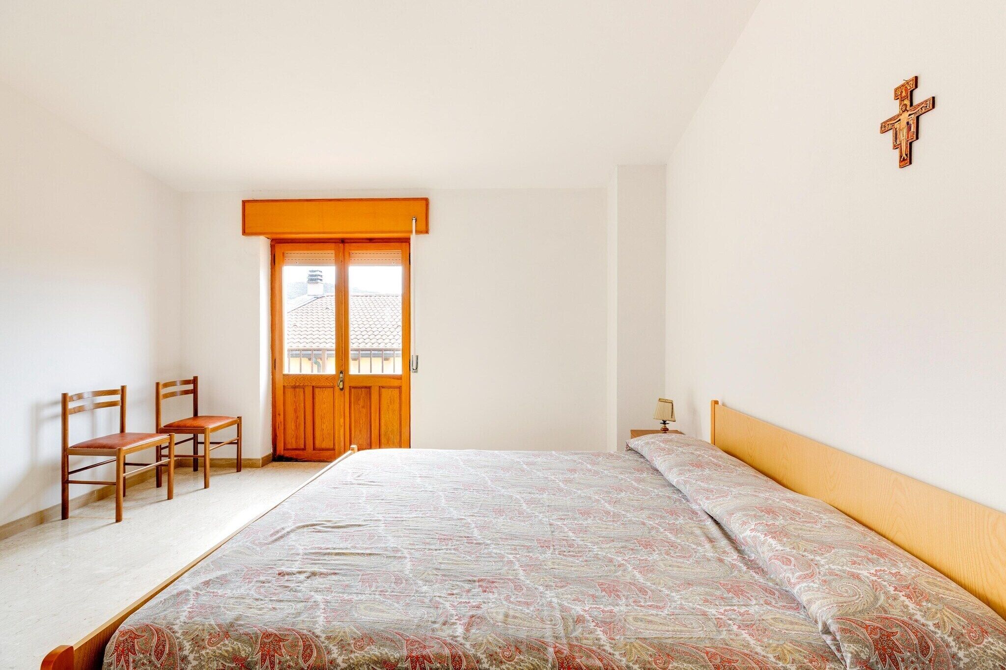 Un dormitorio con cama, dos sillas de madera, una puerta de madera y una cruz en la pared.