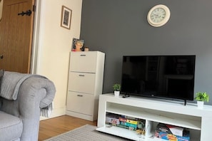 A living room with a grey sofa, a white TV stand with a flat-screen TV, a wooden door, and a small plant on the stand. - Windsor Castle Amazing Cottage | 2-Min Walk | Central & Stylish (Windsor)