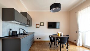 Een moderne keuken met grijze kastjes, een ingebouwde oven en een koffieapparaat. Er is een eethoek met een tafel en stoelen, en een tv aan de muur bevestigd.