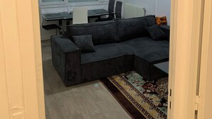 Een ruimtelijke zithoek met een donkergrijze bank, een vloerkleed met patroon en een bureau met een computer.