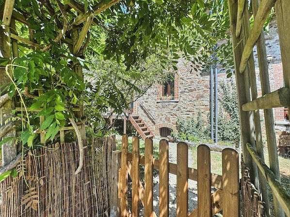 Une clôture rustique en bois agrémentée d’une barrière en bambou, un bâtiment en pierre avec un escalier en briques et une pergola en bois couverte de feuillage verdoyant.