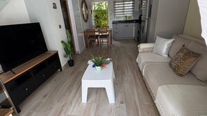 Un salon moderne avec un téléviseur, une table basse blanche, un canapé beige et une cuisine en arrière-plan.