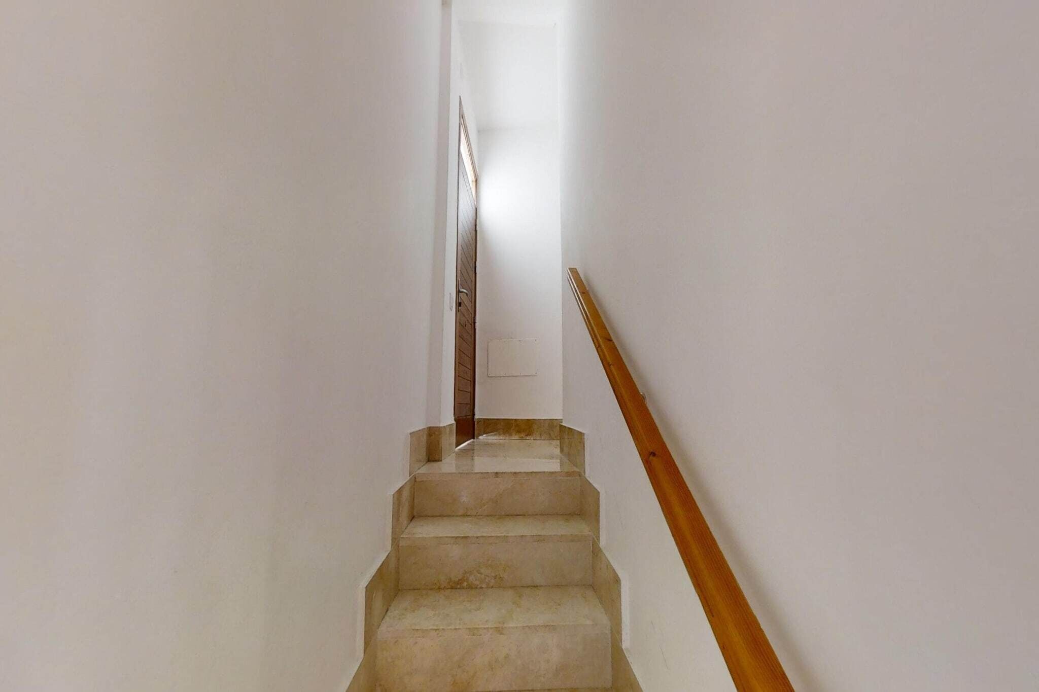 Eine schmale, gut beleuchtete Treppe mit einem Holzgeländer, die zu einer Tür oben führt.