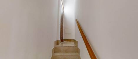 Eine schmale, gut beleuchtete Treppe mit einem Holzgeländer, die zu einer Tür oben führt.