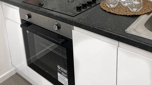 Une cuisine dotée d’un plan de travail noir, d’un four encastré et d’une hotte en acier inoxydable. On y trouve quatre bocaux en verre et quatre verres à vin, posés sur des sets de table en fibres tressées.