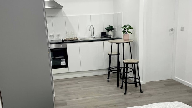 Une chambre compacte comprenant un lit, une petite table et une chaise. À côté de la chambre se trouve une kitchenette équipée d’une cuisinière, d’un évier et de placards.
