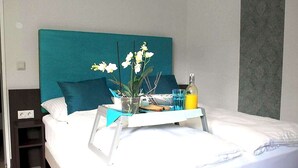 Une chambre d’hôtel avec un lit, une table de chevet, une tête de lit de couleur turquoise, un plateau avec une plante, une bouteille et des verres.