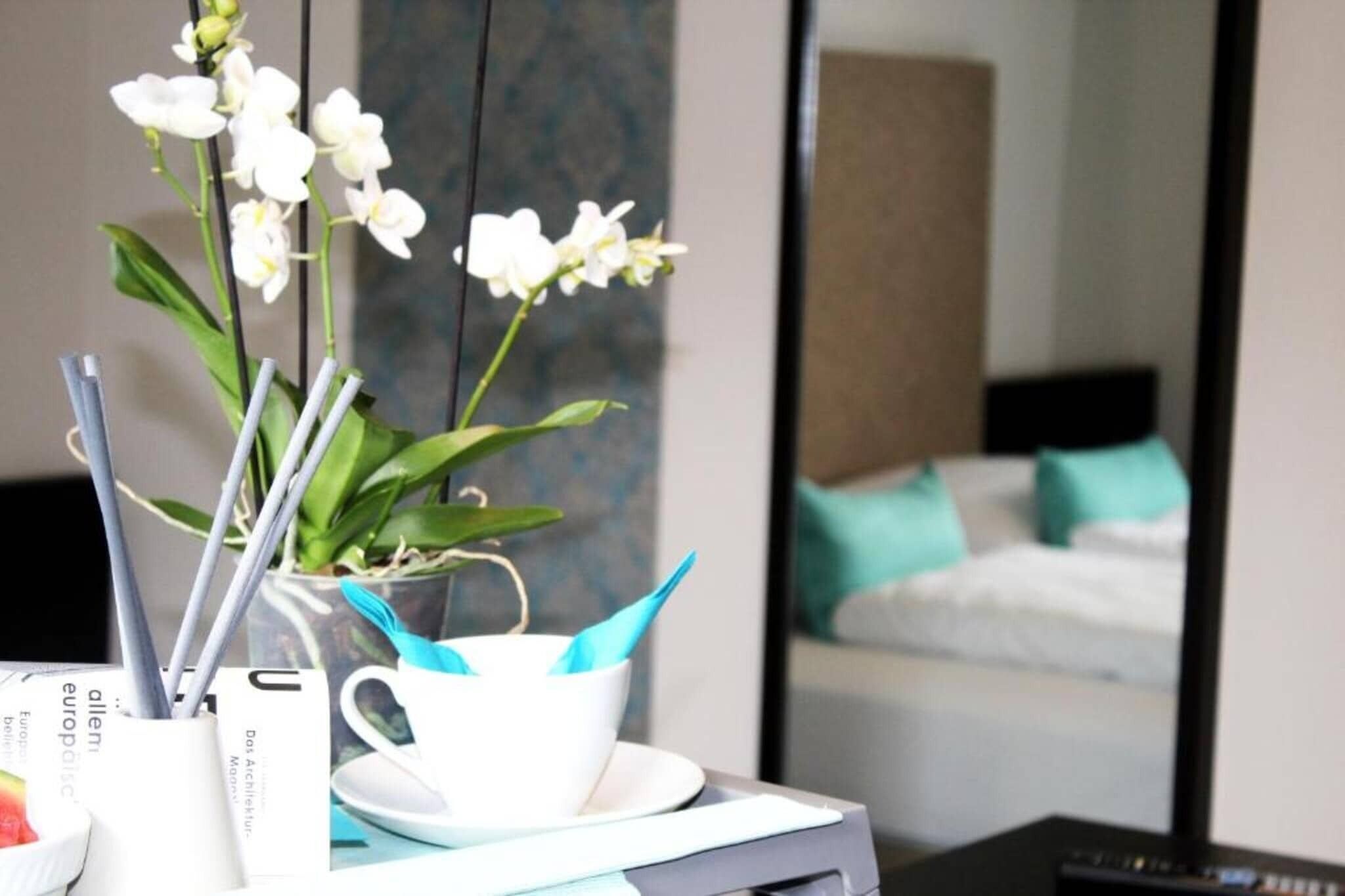 Ein Hotelzimmer mit einem Bett, einem Nachttisch mit Tasse und Untertasse sowie einer Vase mit weißen Blumen.
