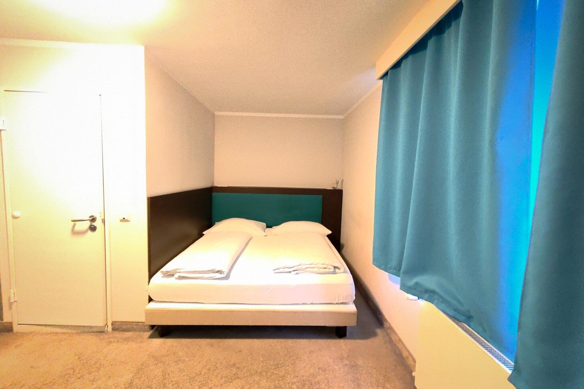 Ein kleines Zimmer mit einem Einzelbett, einem türkisblauen Kopfteil, einer geschlossenen Tür und einem Fenster mit blauen Vorhängen.