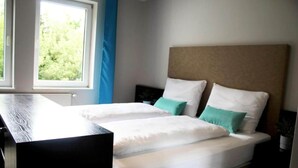 Ein Doppelbett mit weißen und türkisfarbenen Kissen, ein Holz-Kopfteil und ein Fenster mit Blick auf Grünflächen.