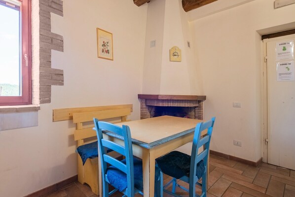 A small dining area with a wooden table and blue chairs, a brick fireplace, and a window with a red frame. - Holiday Home in Sellano with Pool & Sun Terrace (Sellano)