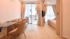 Moderni hotellihuone, jossa on lasipöytä, beigenväriset tuolit, sänky ja parveke, jossa on verhot.