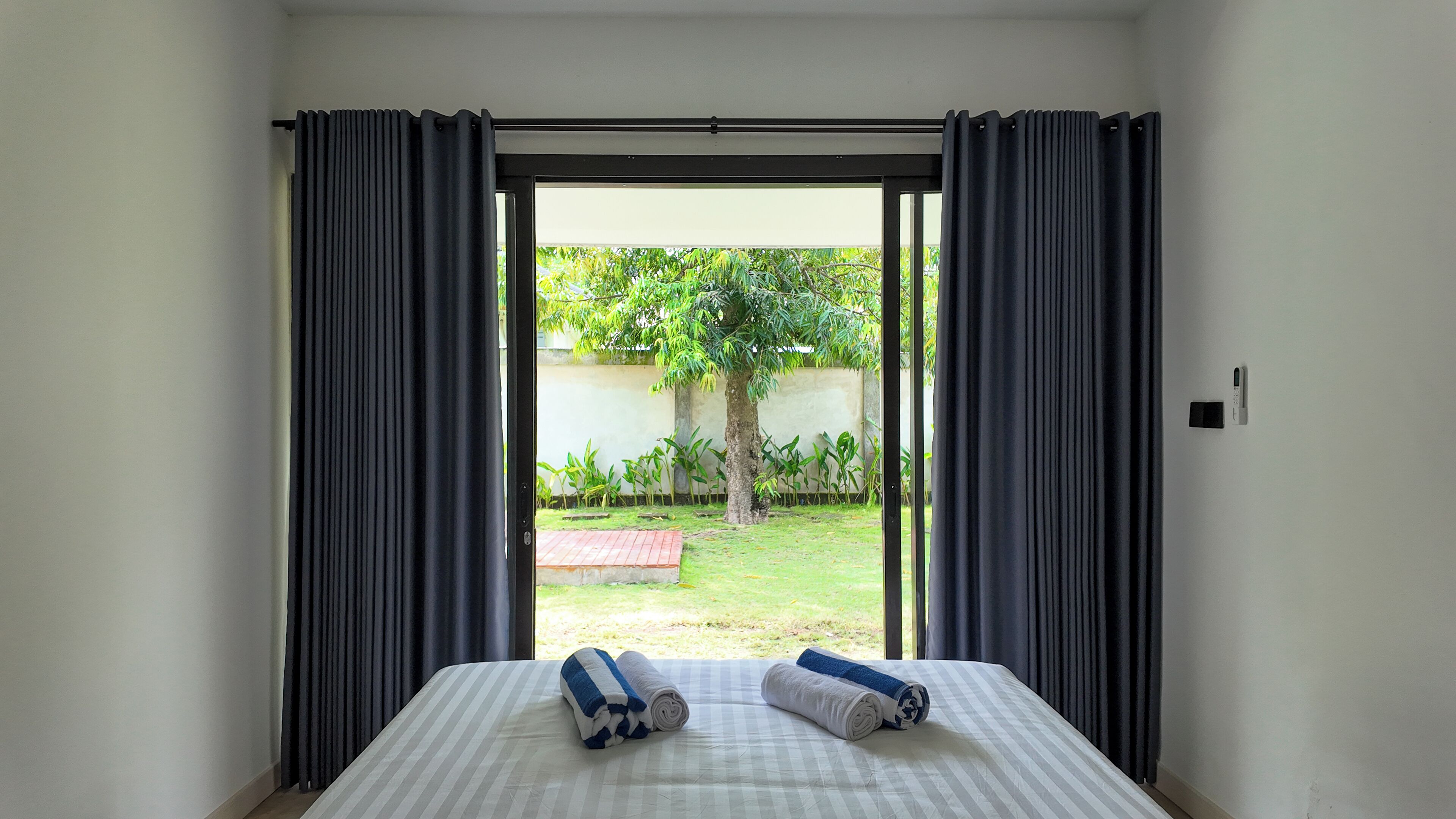 A hotel room with a bed, a view of a garden through an open door, and a small table with a remote control.