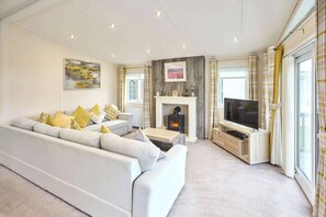 A modern living room with a sofa, fireplace, TV, and large windows. - Host & Stay | The Water Hole Lodge (Pocklington)