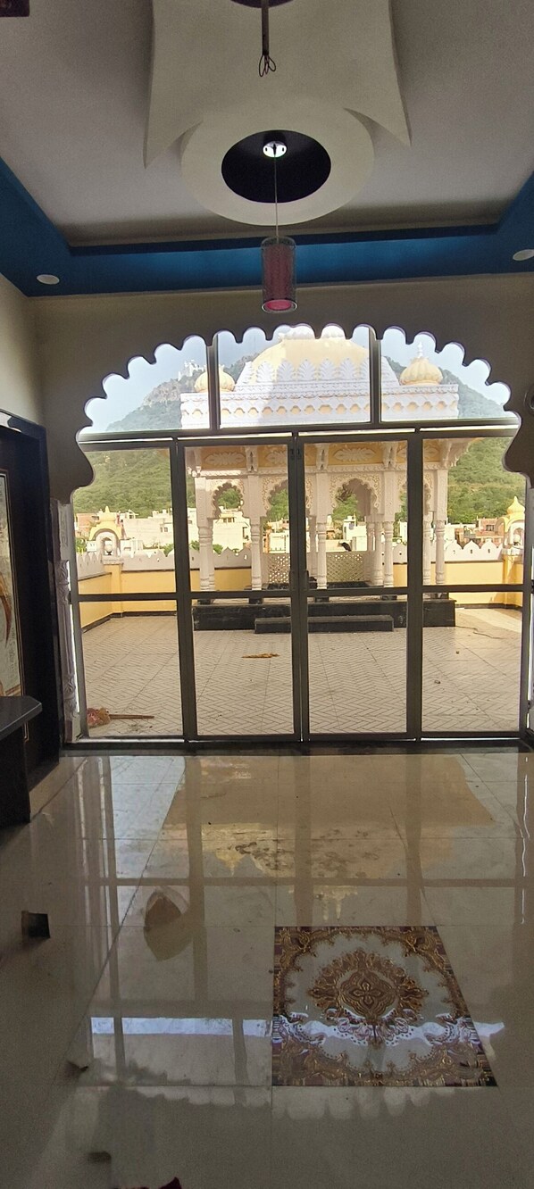 A room with a large glass door leading to an outdoor area with a white structure and a view of a hill. - Shree ji palace  (Udaipur)
