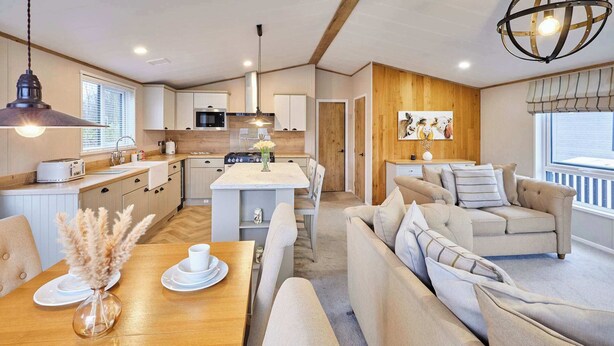A modern kitchen with a microwave, toaster, and wooden cabinets. There is a dining area with a table, chairs, and a vase of dried flowers. A sofa and a lamp are visible in the living area. - Host & Stay | Huckleberry Lodge (Pocklington)