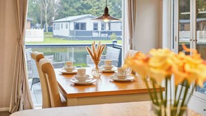 A dining table with white cups and saucers, a vase with dried pampas grass, and a view of a house and water outside a window. - Host & Stay | Huckleberry Lodge (Pocklington)