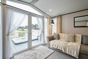 A cozy living room with a sofa, a large window with curtains, a hot tub, and a view of a deck and houses. - Host & Stay | Pebble Lodge (Pocklington)