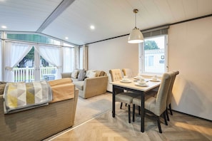 A modern living room with a sofa, a dining table with chairs, and a large window with curtains. - Host & Stay | Elegant Lodge (Pocklington)