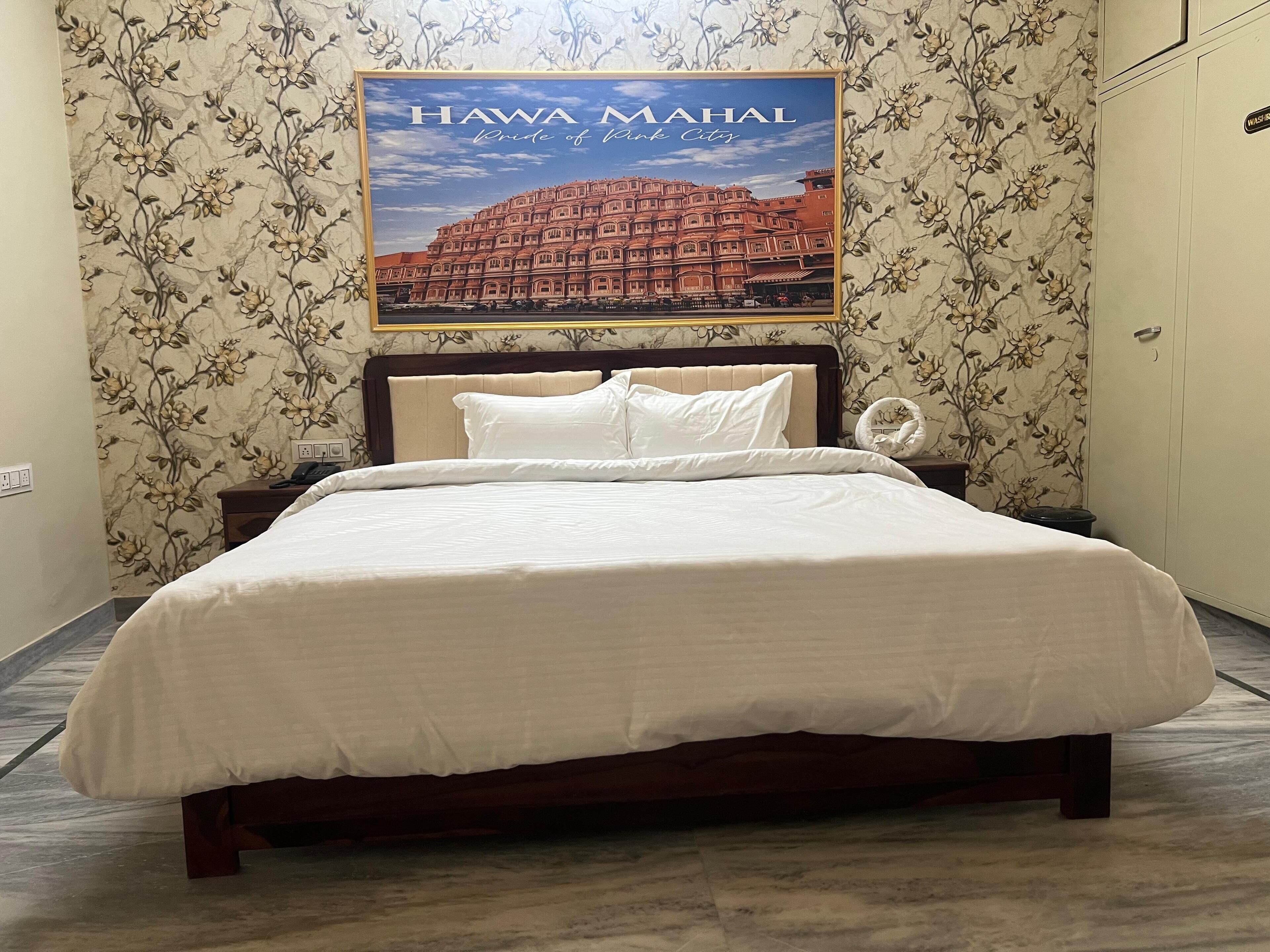 Un lit bien fait, avec une literie blanche, dans une chambre d’hôtel aux murs recouverts d’un papier peint à motifs floraux et orné d’un tableau représentant le Hawa Mahal.