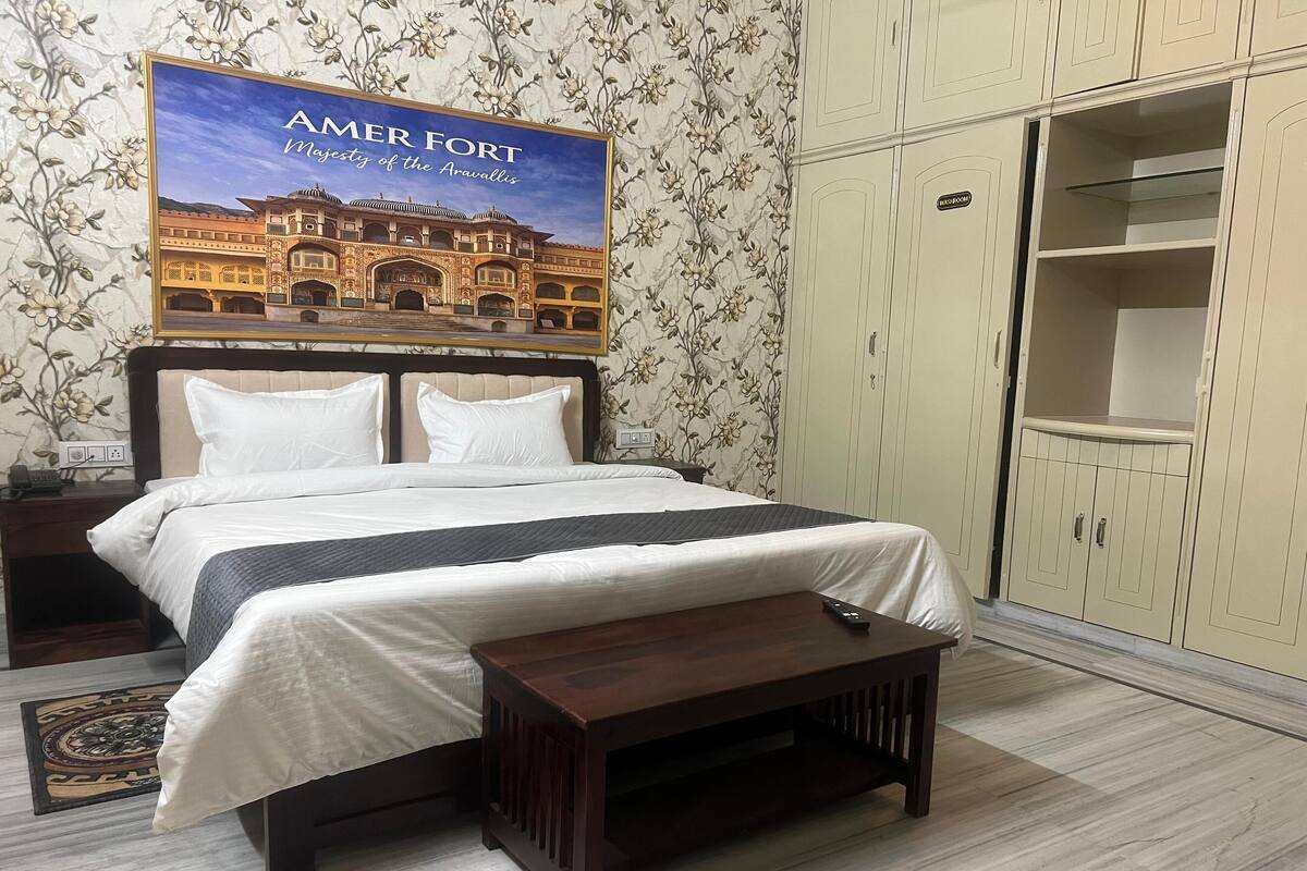 A neatly made bed with a headboard, a bedside table with a lamp, a wooden bench, and a wardrobe with a mirror.