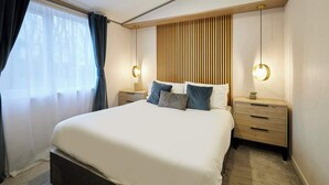Un dormitorio moderno con una cama grande, cabecera de madera, mesitas de noche con lámparas y una ventana con cortinas traslúcidas.