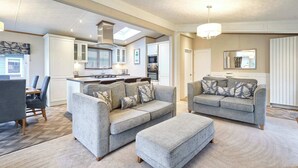 Ein modernes Wohnzimmer mit grauen Sofas und Hocker, ein Essbereich mit Holztisch und Stühlen sowie eine Küche mit weißen Schränken und einem Oberlicht.