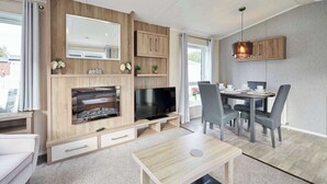 A modern living room with a TV, fireplace, dining area, and a large window. - Host & Stay | Lakeshore Lodge (Pocklington)