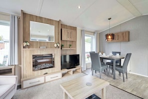 A modern living room with a TV, fireplace, dining area, and a large window. - Host & Stay | Lakeshore Lodge (Pocklington)