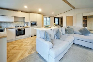 A modern kitchen with built-in appliances, a dining area with a light-colored table, and a grey sofa with cushions. - Host & Stay | Mulberry Lodge (Pocklington)