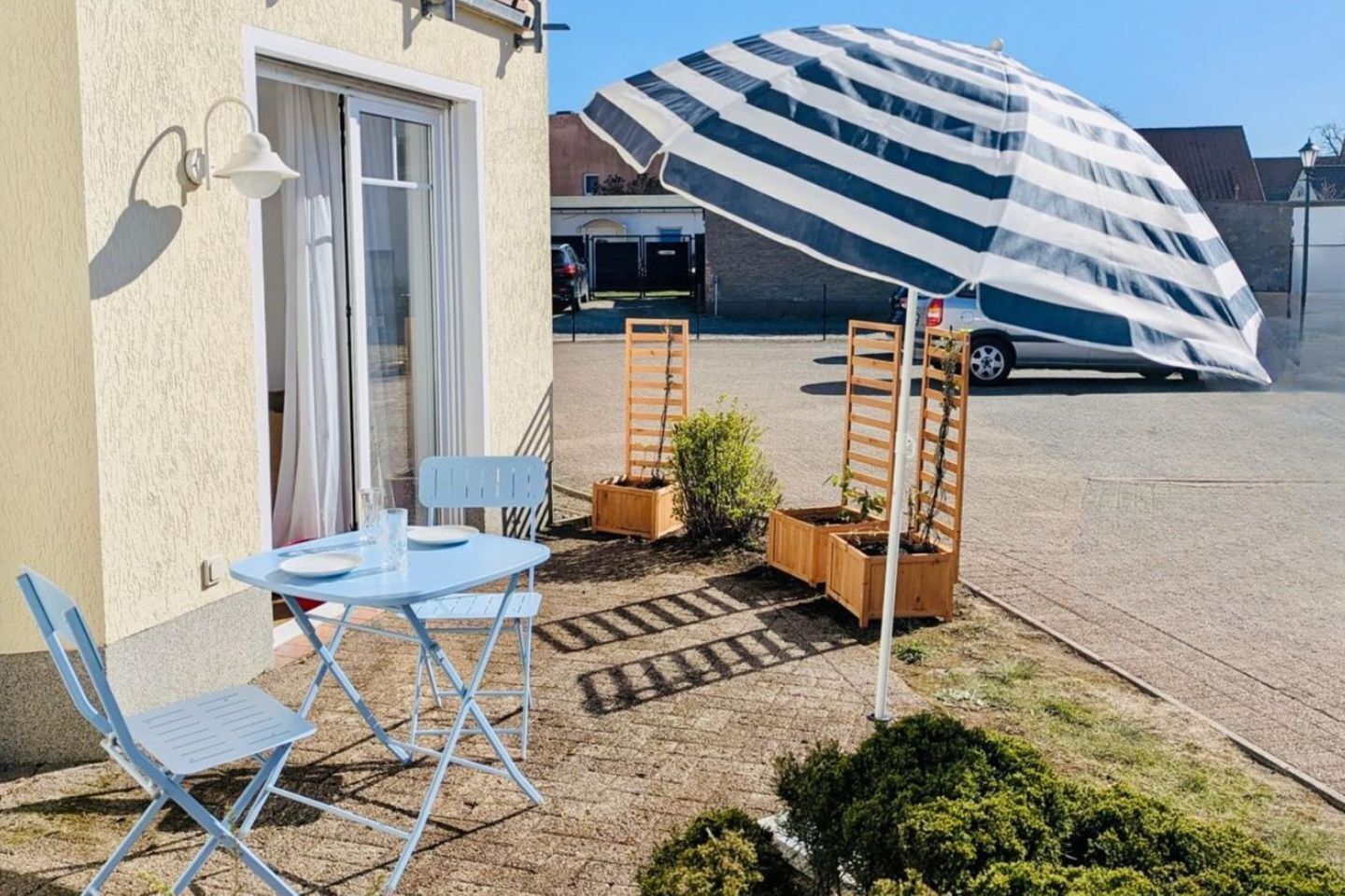 Une terrasse avec une table et des chaises, un petit jardin agrémenté de jardinières en bois et un grand parasol à rayures.
