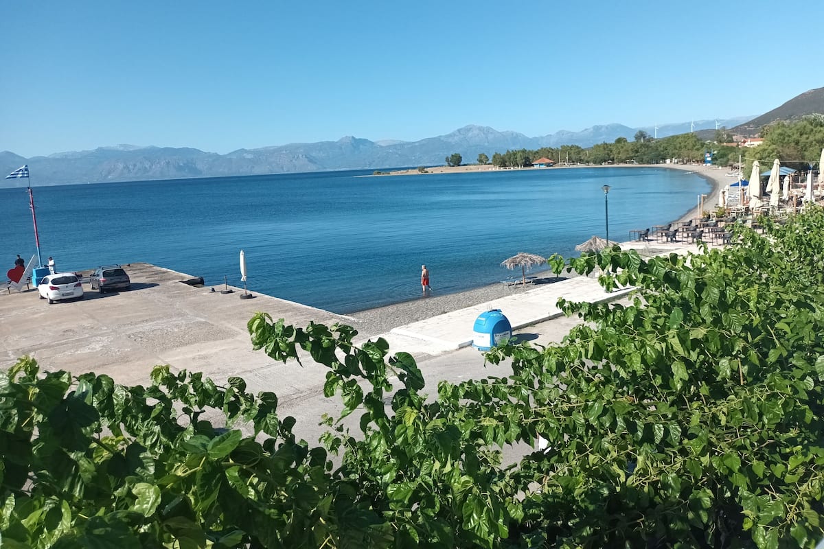 A coastal area with a clear view of the sea, a beach with umbrellas, and a mountainous backdrop.