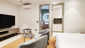 Ett modernt hotellrum med en platt-TV, en soffa, ett litet runt bord, en säng, ett badrum med spegel och handfat samt en luftkonditioneringsanläggning.