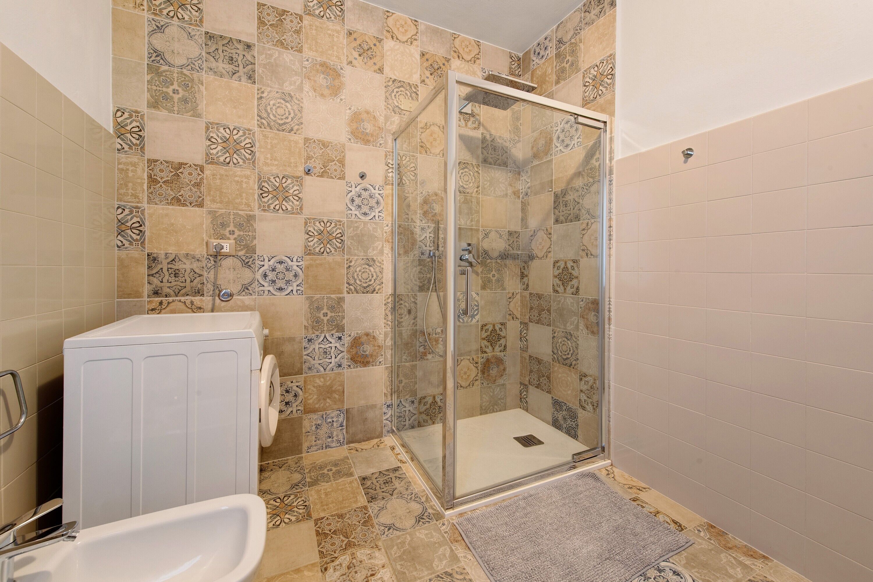 Une salle de bain avec des carreaux décorés, une douche à l’italienne, un lave-linge et un lavabo.