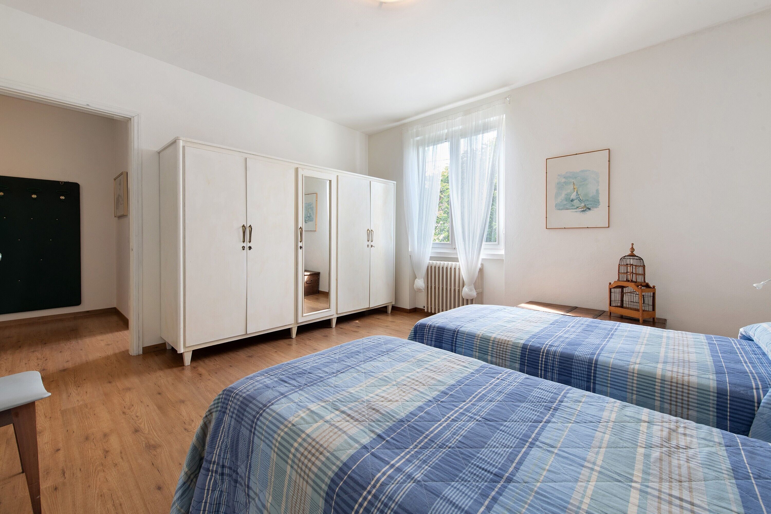A bedroom with two beds, a wardrobe, a radiator, a window with curtains, and a birdcage.