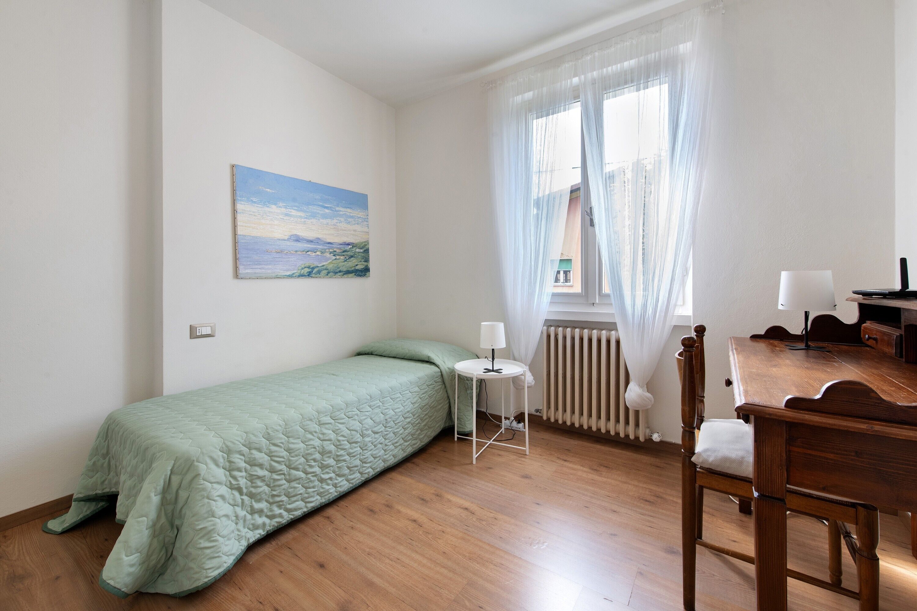 Una stanza piccola arredata con un letto, un tavolo, una sedia, un termosifone e una finestra con tende.