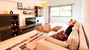 A modern living room with a beige sofa, a glass coffee table, and a patterned rug. There is a TV on a wooden stand, shelves with decorative items, and a window with curtains. - 🌴  Chic & Modern Apartment in the Heart of Hivernage – Pool & Premium Comfort (Marrakech)